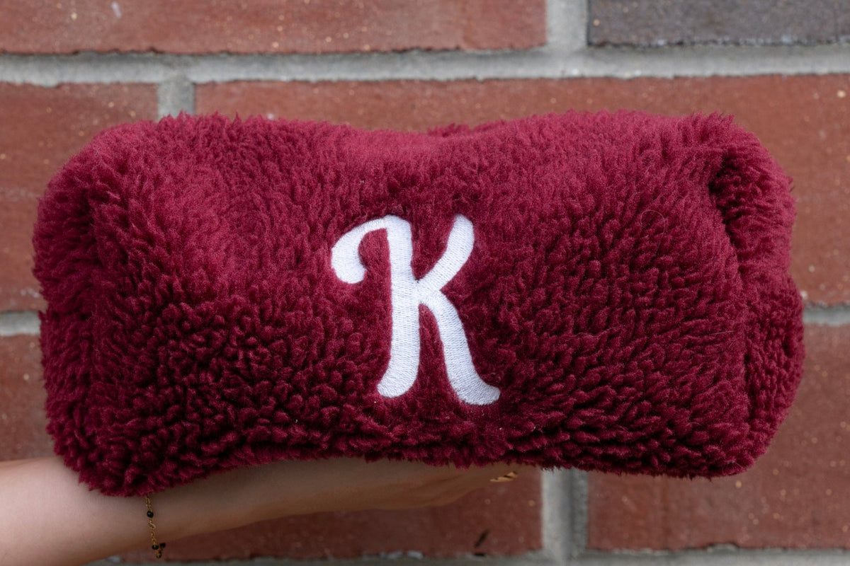 A plush burgundy fleece headband with a white embroidered letter 'K' monogram, held by a hand against a red brick wall background.