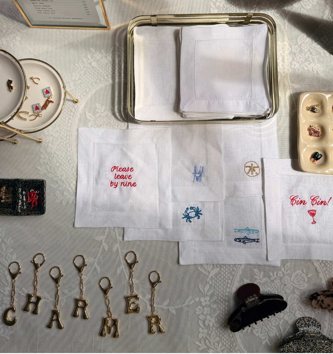 A styled flat-lay of white embroidered napkins on a patterned tablecloth, surrounded by jewelry accessories and small decorative props.